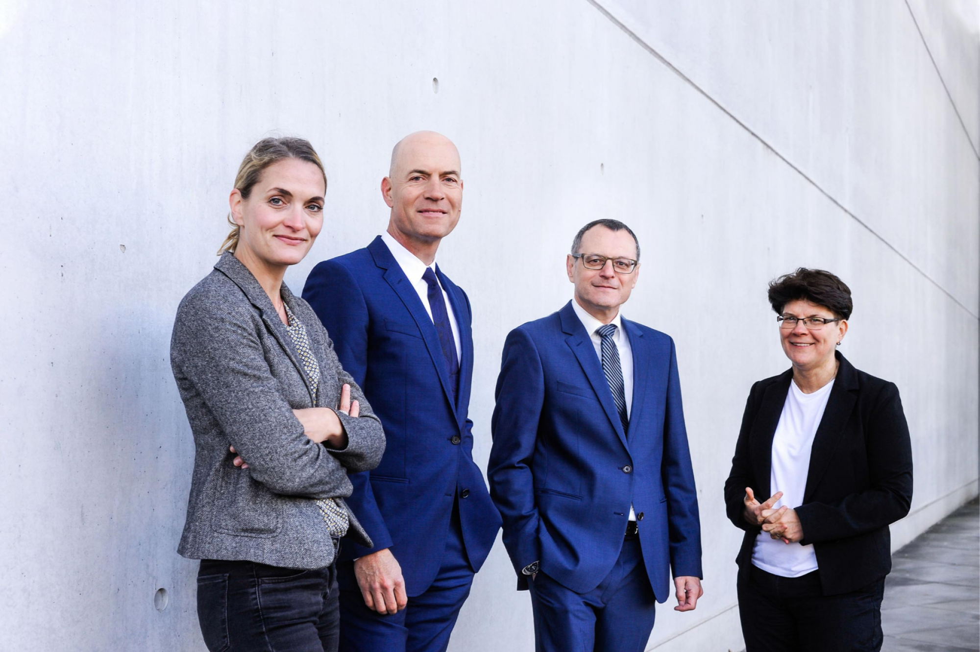 Ein Bild unserer vier Anwälte. Von links nach rechts Dieter Birkeneder, Karin Brandenburger, Jutta Engels und Florian Haußleiter. Sie stehen in Arbeitskleidung mit zuversichtlichem Lächeln vor einem grauen Gebäude mit Glasfassade..