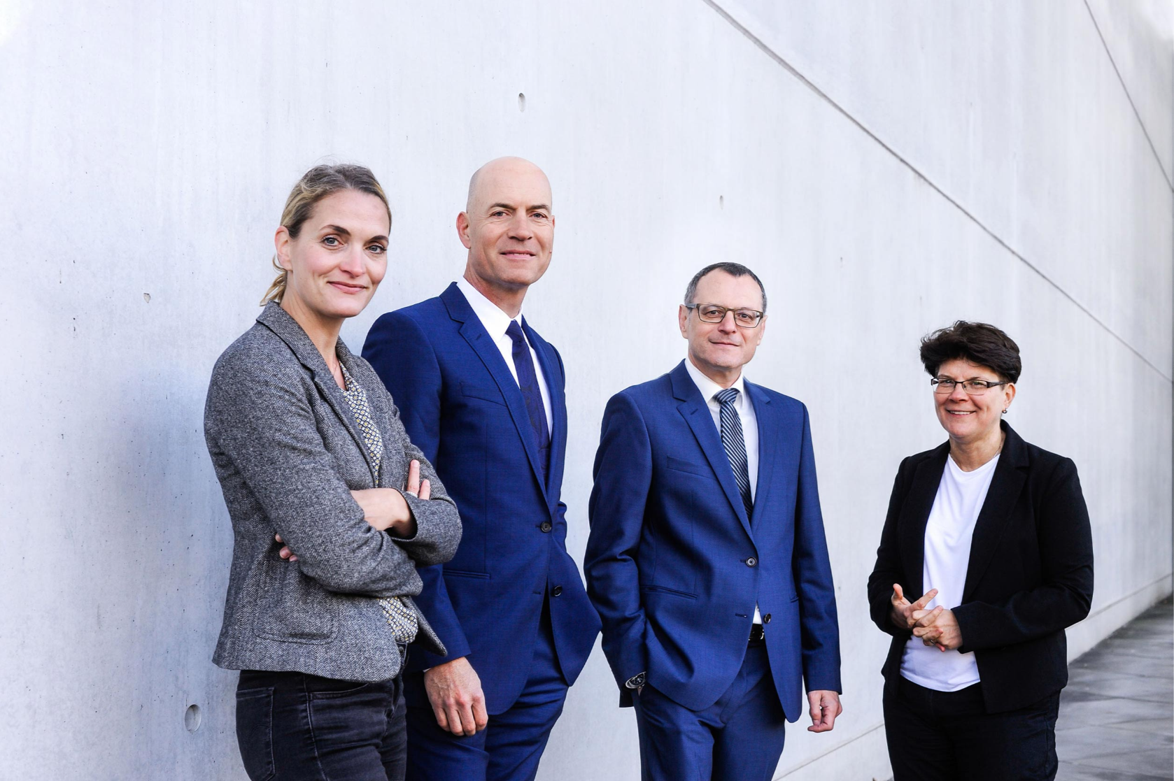 Ein Bild unserer vier Anwälte. Von links nach rechts Jutta Engels, Florian Haußleiter, Dieter Birkeneder und Karin Brandenburger. Sie stehen in Arbeitskleidung mit zuversichtlichem Lächeln vor einer grauen Mauer.