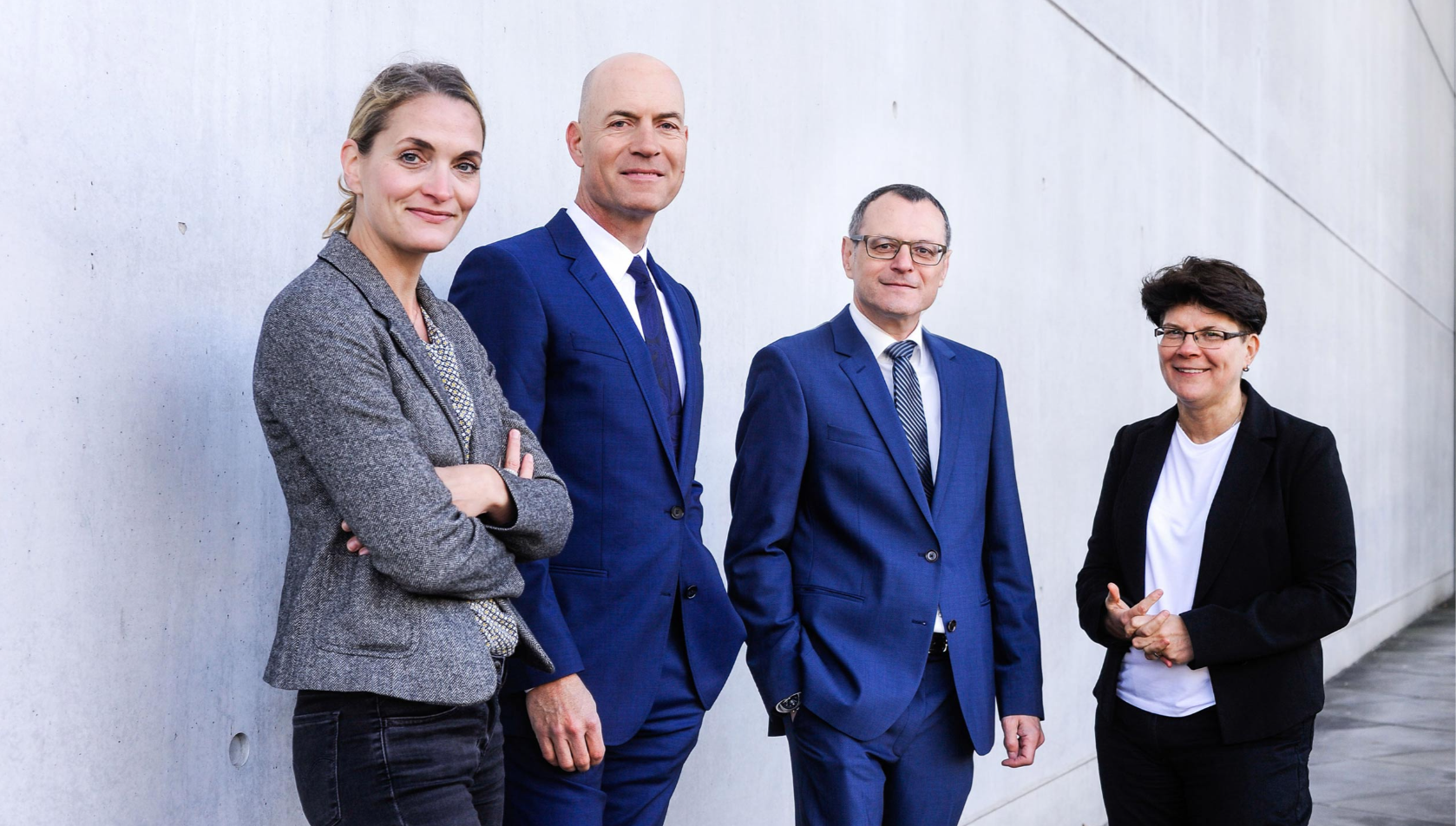 Ein Bild unserer vier Anwälte. Von links nach rechts Jutta Engels, Florian Haußleiter, Dieter Birkeneder und Karin Brandenburger. Sie stehen in Arbeitskleidung mit zuversichtlichem Lächeln vor einer grauen Mauer.
