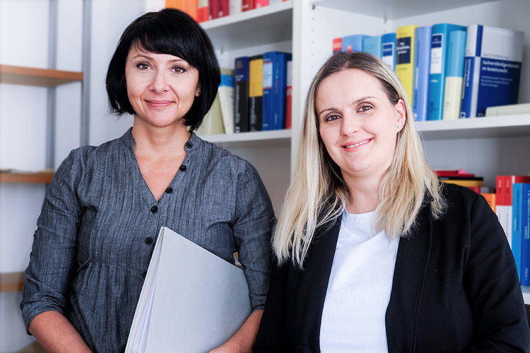 Ein Bild unserer Rechtsanwaltsfachangestellten Doreen Kupfer und Alma Borovac vor einem Bücherregal mit juristischer Lektüre.